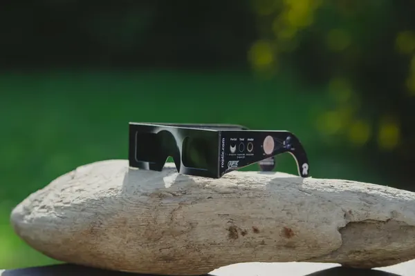 Lunettes d’éclipse ROPTIC posées sur une pierre en extérieur ; sur la branche, les éclipses partielle, totale et annulaire avec leurs dates de 2025 à 2028